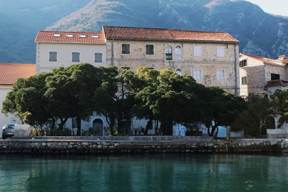 Stone waterfront buildings and trees reflected in the calm water of Boka Bay.