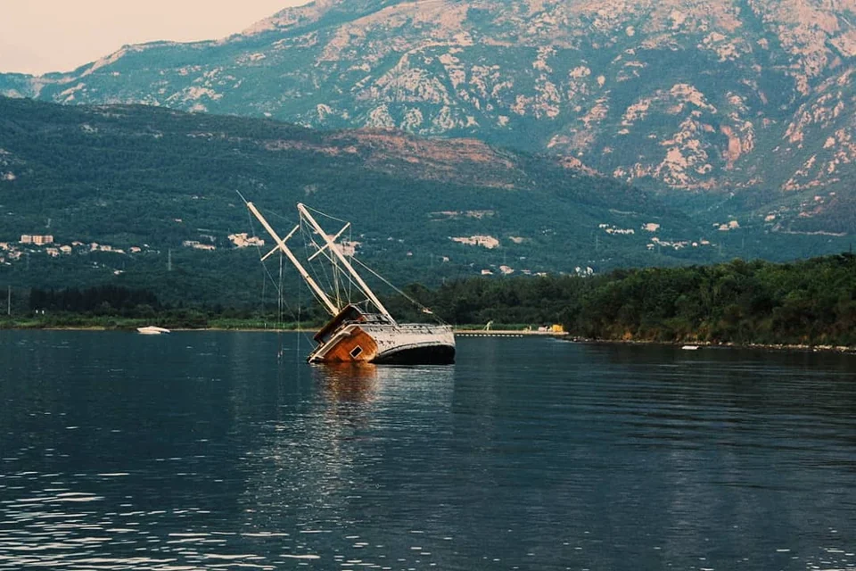 A partly submerged sailing boat near a wooded shore with mountains behind.