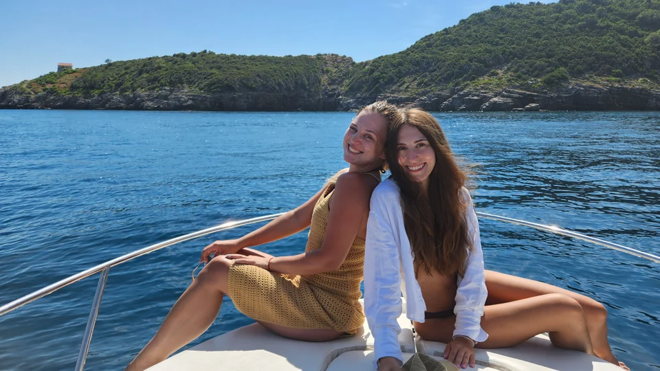 Two guests seated on the bow of Antares 606 beside a wooded coast.