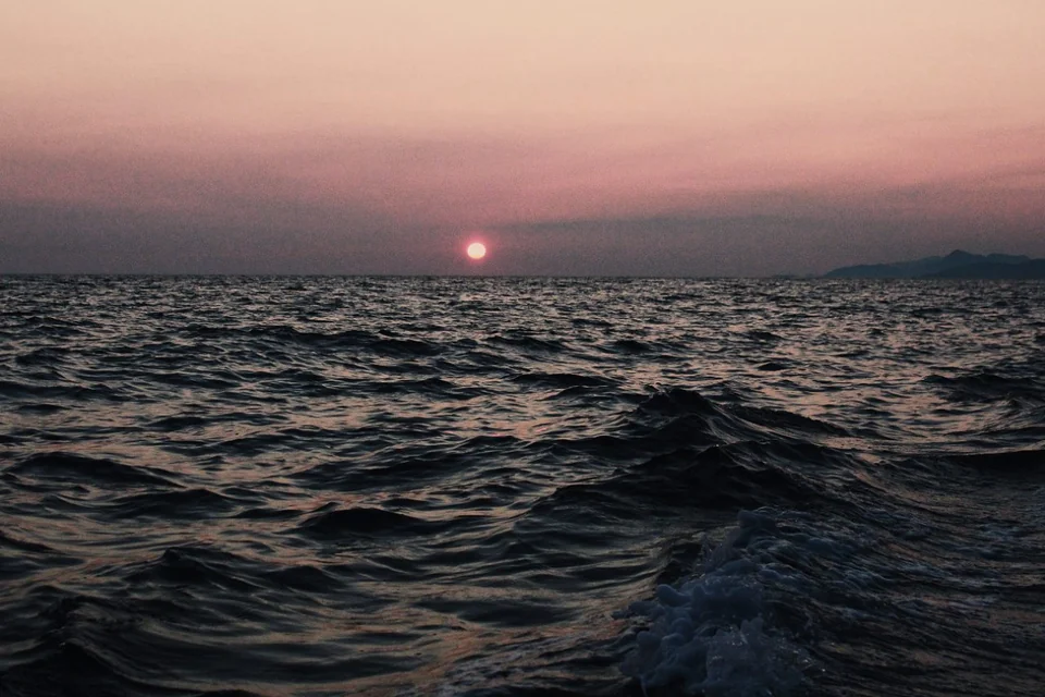 A pink sun low over rippling sea beneath a pale evening sky.