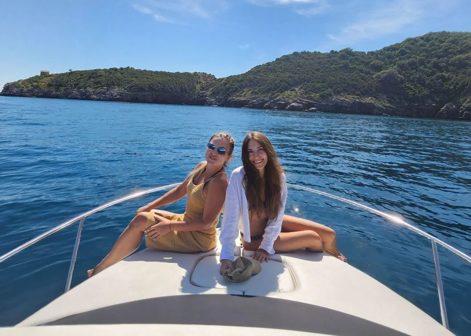 Two guests seated together on the bow with blue water and a wooded coast behind.