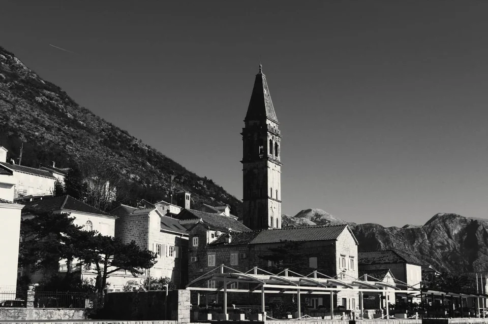 Black-and-white view of Perast's bell tower and stone waterfront buildings.
